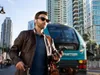 A man is standing on a street with a train in the background, holding a phone and a bag. Produktbild-Vorschau