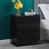 A black high-gloss nightstand with three drawers is placed next to a bed. A vase with flowers and a book with a cup on top are on the nightstand. Produktbild-Vorschau