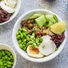A close-up overhead shot of a poke bowl filled with various ingredients, including edamame, rice, egg, cucumber, and pickled onions, presented on a textured gray surface. Produktbild-Vorschau