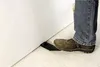 A person's foot wearing a brown shoe is stepping on a black object placed on a light-colored floor next to a white wall. Náhled obrázku produktu
