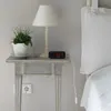 A digital alarm clock sits on a bedside table next to a lamp and a potted plant. Produktbild-Vorschau