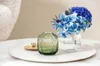 A green glass votive holder and a clear glass vase with blue hydrangeas are displayed on a white tray. Náhled obrázku produktu