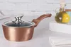 A rose gold saucepan with a glass lid and brown handle is placed on a countertop next to a bottle of olive oil. Náhled obrázku produktu