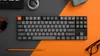A keyboard is placed on a desk divided into gray and orange sections, with a pencil and glasses nearby. Produktbild-Vorschau