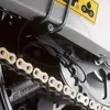 A close-up view of a motorcycle chain and sprocket, with some wiring and a yellow warning sticker visible. Podgląd zdjęcia produktu