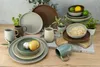 A collection of stoneware dishes and mugs arranged on a wooden surface with some greenery. Produktbild-Vorschau