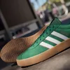 A pair of green Adidas sneakers with white stripes and a gum sole, shown from a side angle on a textured surface. Produktbild-Vorschau