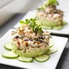 Two plated dishes of food, likely a tuna tartare or similar dish, are presented in a close-up shot. The dishes are layered with avocado and topped with microgreens and what appears to be nori strips. Sliced cucumbers are arranged around the base of the for Produktbild-Vorschau