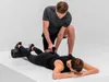 A man is using a massage gun on a woman who is lying on a foam roller. Produktbild-Vorschau