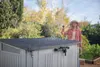 A woman is watering a garden shed with a hose in a backyard setting. Náhled obrázku produktu