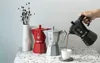 A hand pours coffee from a black moka pot into a white cup, with red and silver moka pots and a vase of dried flowers in the background. Anteprima dell’immagine prodotto
