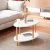 A white oval coffee table with wooden legs is placed on a patterned rug in a living room setting, next to a sofa. A vase with dried flowers and a mug are on the table. Voir les images du produit
