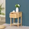 A wooden nightstand with a drawer and a shelf, placed against a blue wall. A vase with flowers and a cup of coffee are on top of the nightstand. Podgląd zdjęcia produktu