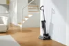 A Tineco wet and dry vacuum cleaner is shown standing upright against a white, ribbed wall in a modern home setting with wooden floors and a staircase in the background. Náhled obrázku produktu