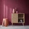 A wooden sideboard with a ribbed sliding door, a vase, and candles on top, placed against a dark pink wall. A pink stool is to the left. Anteprima dell’immagine prodotto