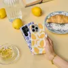 A hand holding a phone case with a lemon pattern, next to lemons, a croissant, and tea. Náhled obrázku produktu
