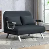 A dark gray sofa bed with two throw pillows is shown in a room with a patterned rug and a window in the background. Náhled obrázku produktu