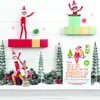 A festive display featuring Elf on the Shelf toys, miniature Christmas trees, and a book, arranged on a shelf with snow and festive decorations. Produktbild-Vorschau