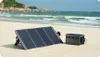 A portable solar power station and solar panel are set up on a sandy beach with the ocean and rocky hills in the background. Náhled obrázku produktu