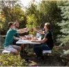 Three men are toasting with bottles at a picnic table outdoors. Produktbild-Vorschau