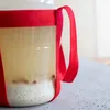 A close-up of a glass container holding a light-colored liquid with some sediment and bubbles at the bottom, wrapped in a red ribbon. Produktbild-Vorschau