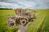 A wooden model of a combine harvester is held in a person's hand in a field of green crops. Podgląd zdjęcia produktu