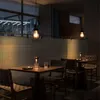 A dimly lit restaurant or cafe setting with tables and chairs, illuminated by pendant lights and table lamps. Produktbild-Vorschau