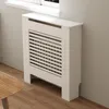 A white radiator cover is placed against a light gray wall, next to a window with a white windowsill. A wooden coffee table is partially visible in the foreground. Anteprima dell’immagine prodotto