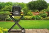 A black grill stand with an open grill on top, set up on a paved patio in a lush green garden. Náhľad obrázku produktu
