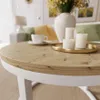 A close-up of a round coffee table with a wood-grain top and white base, set in a living room with a sofa and lamp in the background. Náhled obrázku produktu