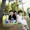 Three children are sitting on a nest swing in a park-like setting. Produktbild-Vorschau