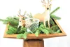 A decorative arrangement of two deer figurines, a glass jar, and evergreen branches in a wooden tray. Produktbild-Vorschau