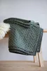 A folded green knitted blanket is placed on a wooden bench, with a fluffy white rug to the left. Produktbild-Vorschau
