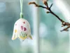 A decorative bell ornament with floral and butterfly designs hangs from a ribbon, set against a blurred background of a window and a tree branch. Produktbild-Vorschau