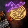 A Halloween-themed neon sign of a pumpkin wearing a witch's hat, placed on a dark, textured surface with fallen leaves and a small chalkboard with the word 'Boo'. Produktbild-Vorschau