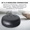 A black portable speaker is shown on a light gray surface, with a blurred background of a hand holding a phone. Produktbild-Vorschau
