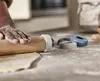 A person's hands are using a rolling pin to flatten dough on a floured surface. Several ring-shaped guides for the rolling pin are visible nearby. Produktbild-Vorschau