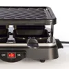 Close-up view of a black electric raclette grill with a ribbed top surface and small pans underneath. Produktbild-Vorschau
