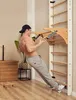 A muscular man is performing a pull-up exercise on a wooden wall-mounted gym apparatus in a room. Produktbild-Vorschau