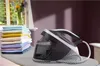 A steam iron station and a stack of folded laundry are displayed on an ironing board next to a window with flowers. Voir les images du produit