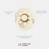 A cream-colored pacifier with a decorative shield and a gold ring around the nipple, shown against a light gray background with descriptive text labels pointing to different features. Produktbild-Vorschau