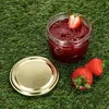 A jar of strawberry jam with strawberries and its lid on a grassy surface. Produktbild-Vorschau