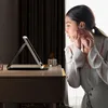 A woman is applying makeup in front of a lighted mirror on a vanity. Produktbild-Vorschau