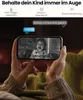 A person holding a smartphone displaying a baby monitor feed, with text overlays indicating features. Produktbild-Vorschau