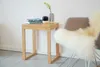 A small wooden stool with a book and vase on top, next to a chair with a sheepskin rug. Produktbild-Vorschau
