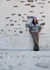 A person leans against a textured stone wall, wearing a knitted vest and hat. Produktbild-Vorschau