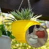 A yellow hanging planter with a spider plant is shown outdoors, with a circular inset showing a person's feet standing on two different colored plant pots. Podgląd zdjęcia produktu