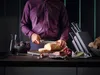 A person is slicing bread on a cutting board, with various food items and a knife block on a counter. Voir les images du produit