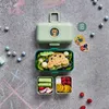 A green MONBENTO lunchbox is open and filled with food, placed on a grey, chalk-drawn surface. Produktbild-Vorschau
