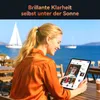 A woman is sitting at a table outdoors, looking at a tablet with a scenic background of the sea and sailboats. Produktbild-Vorschau